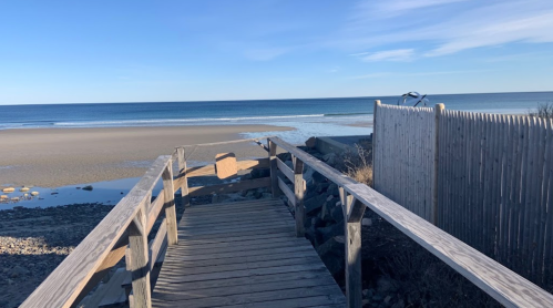 A wooden walkway leads to a calm beach with gentle waves and a clear blue sky. A fence is visible on the right.