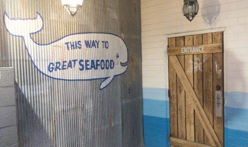 A corrugated metal wall with a whale mural and a wooden door labeled "Entrance" leading to a seafood restaurant.