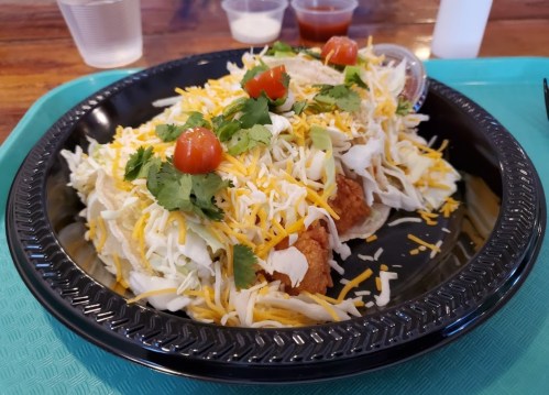 A plate of tacos topped with shredded lettuce, cheese, cilantro, and tomatoes, served with sauces on the side.