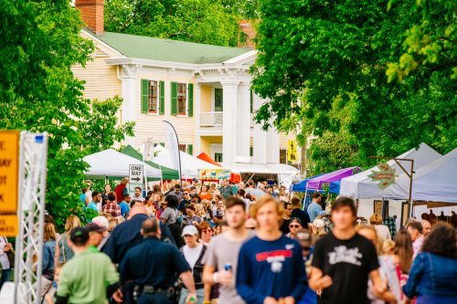 A bustling street fair with crowds, tents, and a historic building in the background, surrounded by greenery.