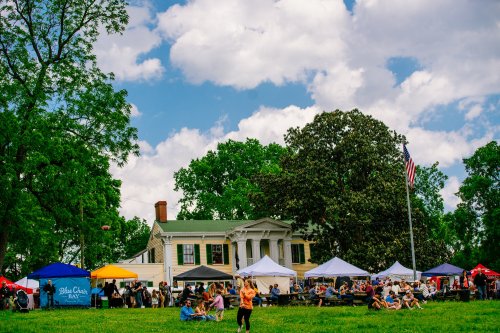 A lively outdoor event with tents and people gathered in front of a historic house under a blue sky with clouds.