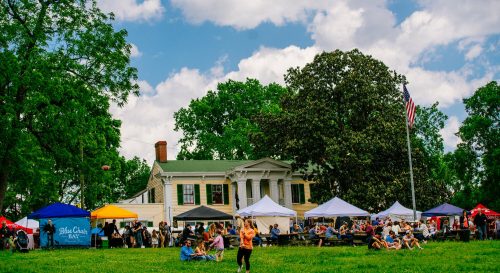 A lively outdoor event with tents and people gathered in front of a historic building under a blue sky with clouds.