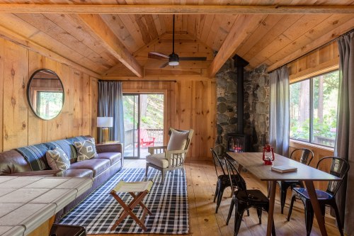 Cozy cabin interior with wooden walls, a leather sofa, dining table, and large windows overlooking a forested area.