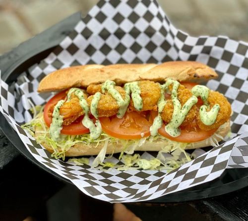 A sub sandwich filled with shrimp, lettuce, and tomatoes, drizzled with green sauce, served in a checkered paper basket.
