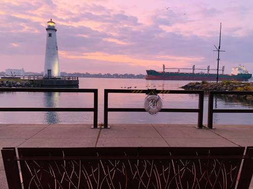A serene waterfront view featuring a lighthouse, a ship, and a bench at sunset.