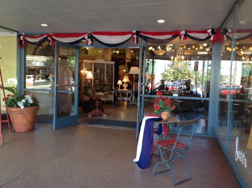 A storefront with large glass doors, decorated with red, white, and blue bunting, and outdoor seating with potted plants.
