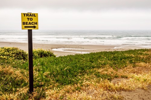 A yellow sign points left, indicating a trail to the beach, with ocean waves and sandy shore in the background.