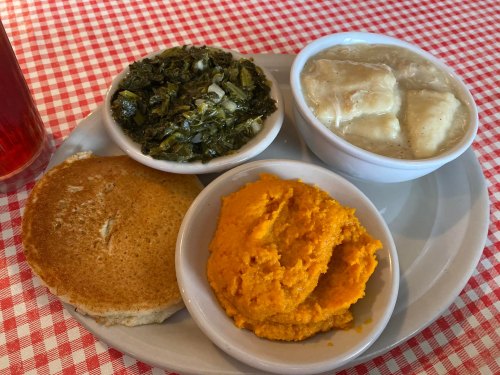 A plate with cornbread, collard greens, dumplings in broth, and sweet potato mash on a checkered tablecloth.