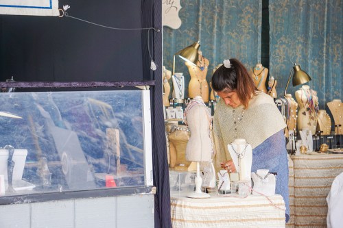 A woman works on jewelry at a display, surrounded by mannequins and various accessories in a market setting.