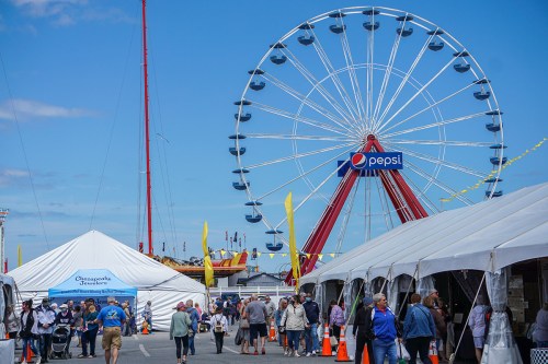 A busy fairground scene with a large Ferris wheel, tents, and crowds of people enjoying the festivities.