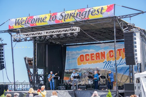 A band performs on stage at Springfest in Ocean City, Maryland, with a colorful banner overhead and an audience in view.