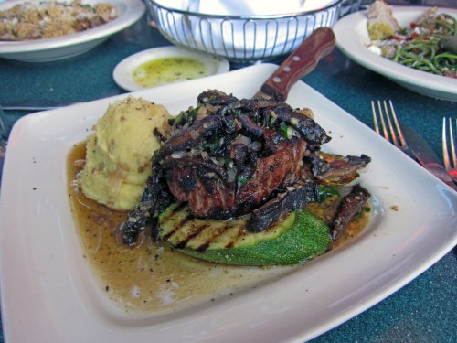 A plated dish featuring grilled steak on zucchini, topped with mushrooms, served with mashed potatoes and sauce.