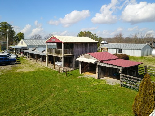 Aerial view of a rustic barn complex with multiple buildings and parked cars on a grassy area under a blue sky.
