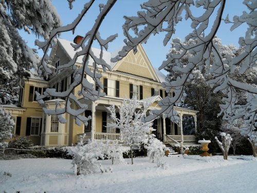 A snow-covered Victorian house surrounded by trees and a winter landscape, creating a serene, picturesque scene.