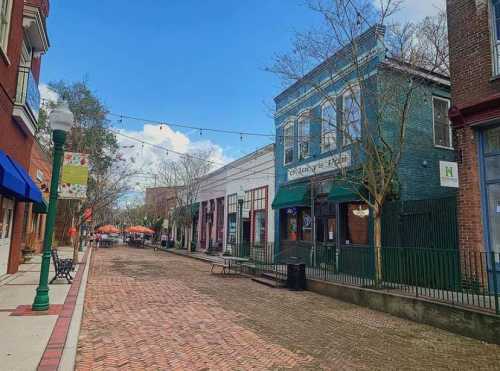 A charming brick street lined with shops and trees, under a blue sky with scattered clouds. Outdoor seating is visible.