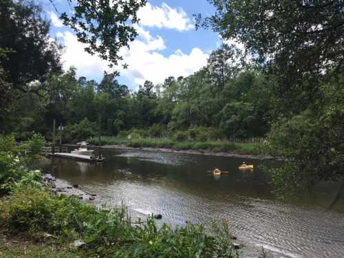 A serene river scene with kayakers, lush greenery, and a small dock under a partly cloudy sky.