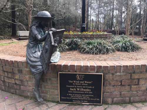 A bronze statue of a girl painting, with a plaque honoring Jack Wilbanks in a landscaped park setting.