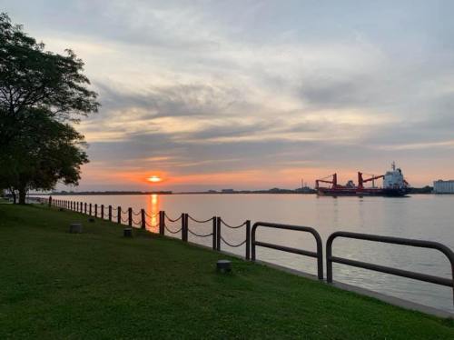A serene sunset over a calm river, with a ship docked and trees lining the grassy shore.