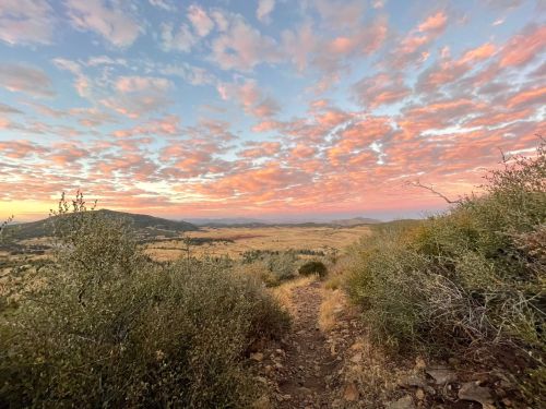 A scenic view of a landscape at sunset, featuring rolling hills, sparse vegetation, and a colorful sky filled with clouds.