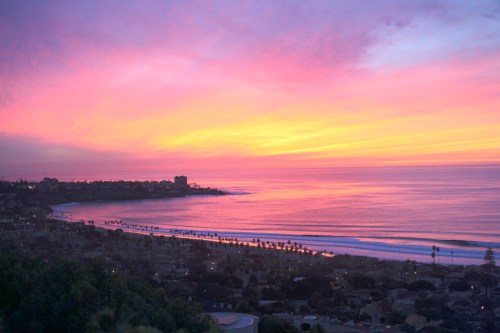 A vibrant sunset over a calm ocean, with pink and purple hues reflecting on the water and a coastal town in the foreground.