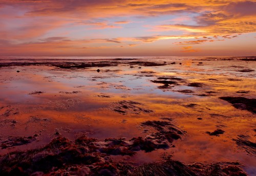 A vibrant sunset reflects on a calm ocean, with colorful clouds and wet sand creating a serene coastal scene.