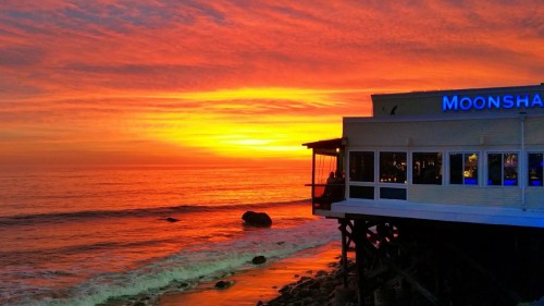 A vibrant sunset over the ocean, with a restaurant on stilts silhouetted against the colorful sky.