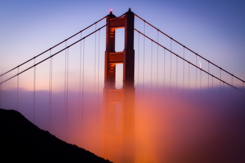 Golden Gate Bridge partially shrouded in fog at sunset, with warm hues illuminating the structure.