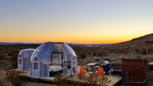 A transparent dome structure in a desert landscape at sunset, with outdoor seating and mountains in the background.