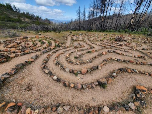 A winding stone labyrinth set in a grassy area, surrounded by charred trees and a distant view of hills under a cloudy sky.