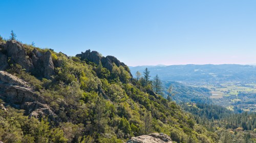 A scenic view of lush green hills and rocky outcrops under a clear blue sky, overlooking a valley below.