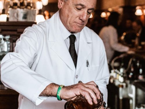 A bartender in a white coat pours a drink at a bar, focused on his task amidst a lively atmosphere.