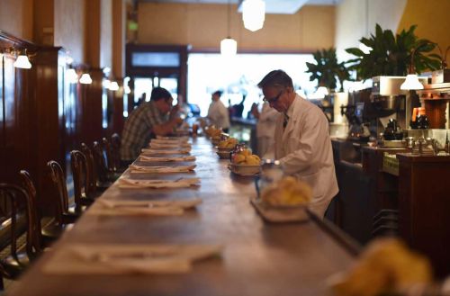 A vintage diner scene with a long wooden counter, patrons seated, and a server in a white coat preparing food.