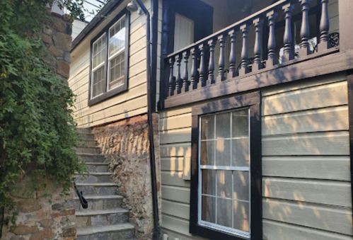 A staircase leads up to a house with a balcony, featuring wooden railings and light-colored siding.