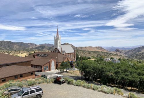 A scenic view of a church and buildings nestled in a mountainous landscape under a blue sky with wispy clouds.