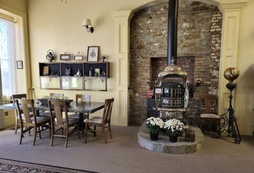 Cozy room with a wooden dining table, chairs, and a vintage stove against a stone wall, decorated with flowers.