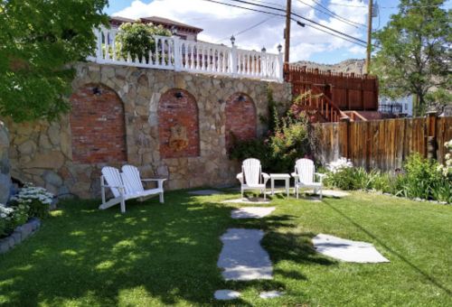 A serene garden with stone walls, three white chairs, and a grassy area surrounded by flowers and trees.