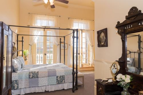Cozy bedroom with a four-poster bed, large windows, and vintage decor, featuring a mirror and floral arrangement.