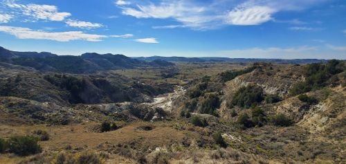 A panoramic view of rolling hills and valleys under a clear blue sky, with sparse vegetation and distant mountains.