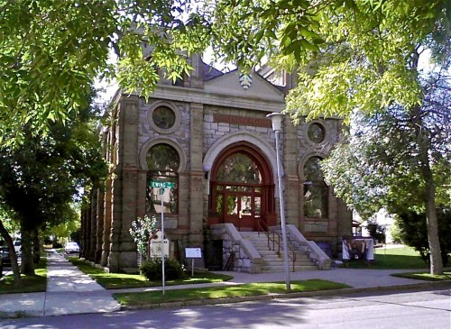 Historic stone building with large arched entrance, surrounded by trees and greenery on a sunny day.