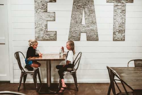 Two young girls sit at a table in a cozy restaurant, enjoying drinks and chatting, with a large "EAT" sign on the wall.