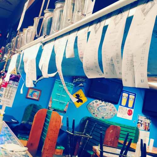 A colorful bar interior with hanging receipts, jars on a shelf, and wooden chairs, creating a lively atmosphere.