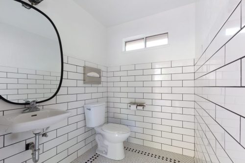 A modern bathroom featuring white subway tile, a round mirror, a sink, and a toilet with a small window above.