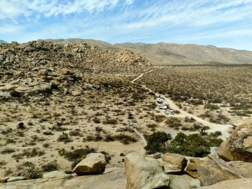 A rocky landscape with sparse vegetation and a dirt road leading to parked vehicles in the distance under a blue sky.