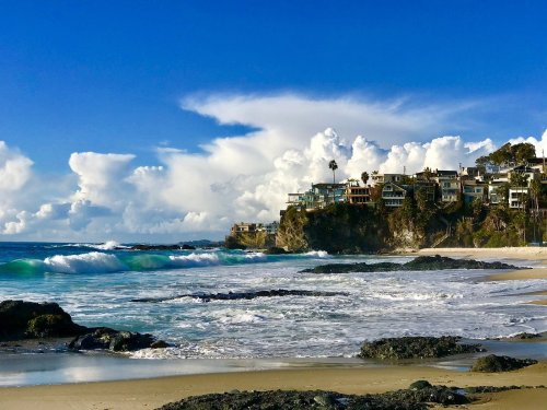 A scenic beach view with waves crashing on rocks, cliffs, and houses under a bright blue sky with fluffy clouds.