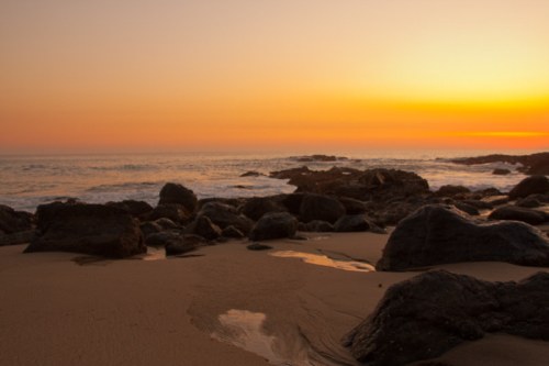 Sunset over a rocky beach, with gentle waves lapping at the shore and a warm orange sky.