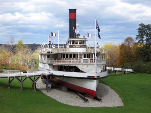 A historic steamboat docked on land, surrounded by green grass and trees, with flags flying on a cloudy day.