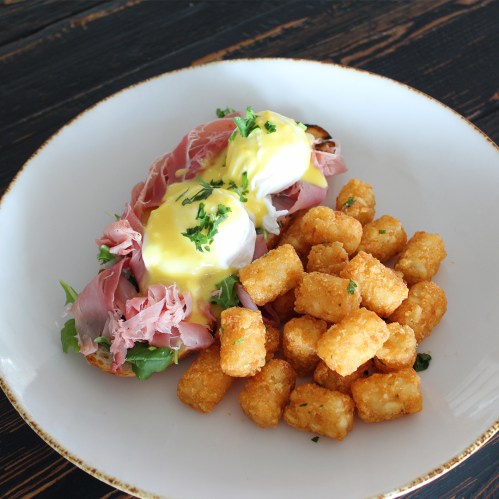 A plate with poached eggs on ham and greens, served with crispy tater tots, garnished with parsley.