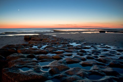 A serene beach at sunset, featuring rocky formations and calm waters reflecting the colorful sky.