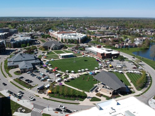 Aerial view of a community event with tents and activities on a green space, surrounded by buildings and trees.