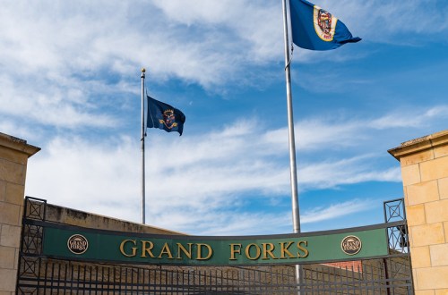 Entrance sign for Grand Forks with flags flying against a blue sky.